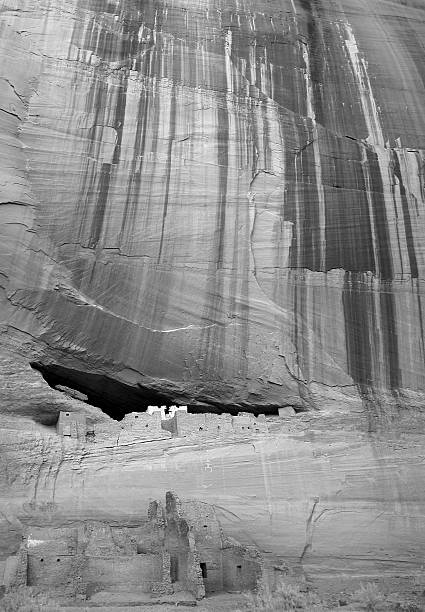 Massive red vertical canyon walls dominate a tiny cliff ruin in center of photo as seen from Junction Overlook in Canyon de Chelly National Monument.