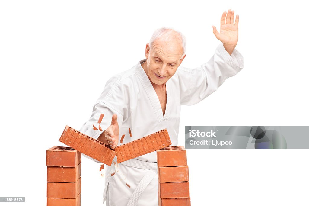 Senior breaking a brick with his bare hand Studio shot of a senior man in a white kimono breaking a brick with his bare hand isolated on white background Breaking Stock Photo