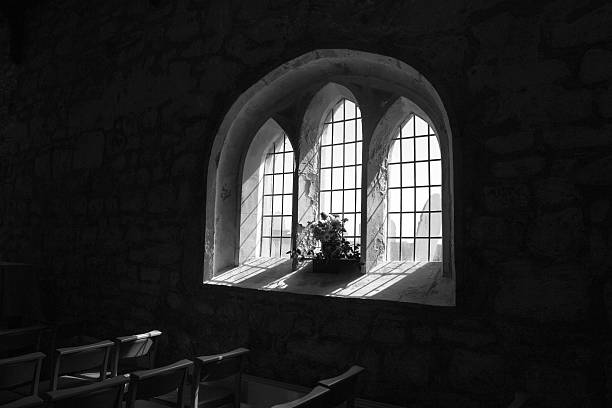 Bright sunlight glowing through a simple stained glass window in the darkness inside the church of St Peter and St Paul in Shernborne, a tiny village in Norfolk, Eastern England. The current reconstructed church was built in 1898 and paid for by King Edward VII but the site on which it stands dates back to the 7th century. The font within the church is Norman and much of the stonework within the church is thought to be original.