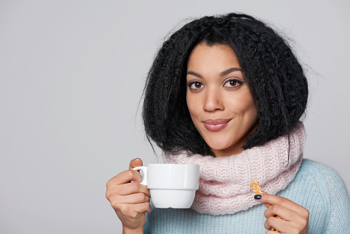 Smiling mixed race african american - caucasian girl wearing warm knitted sweater and scarf drinking hot drink from a cup with a biscuit, over gray background
