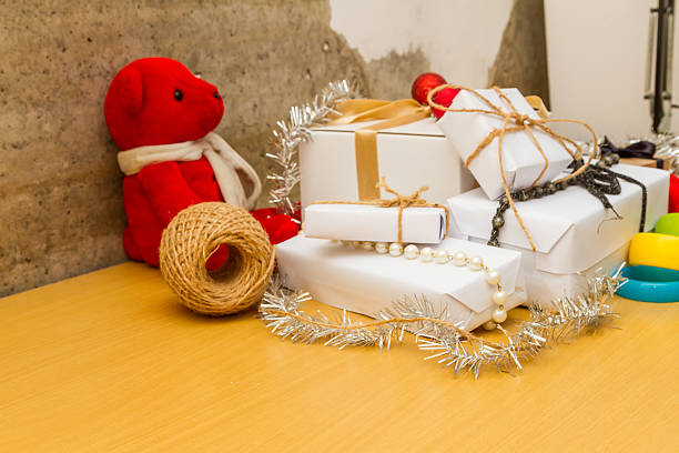 A middle-aged man in casual gray sweatpants and white socks sits cross-legged on a gray carpeted living room floor surrounded by holiday wrapping supplies. He appears relaxed during gift-wrapping preparations, with rolls and cut sheets of white paper printed with brown reindeer patterns nearby, a gold-colored tape dispenser on the floor, and a wooden side table in the background. The indoor setting features neutral tones and soft lighting, evoking a cozy winter atmosphere. This image suits commercial uses in holiday lifestyle articles, seasonal marketing for home goods, family tradition features in media, or e-commerce promotions for wrapping accessories and festive decor.