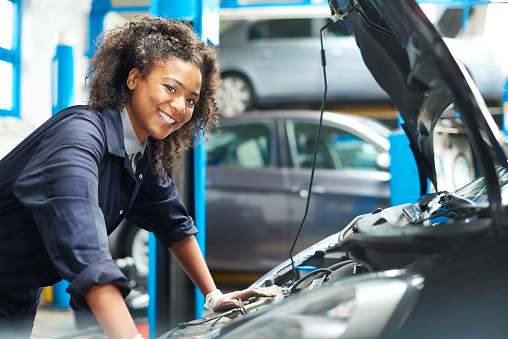 A female mechanic is working under a bonnet of a car in a garage repair shop. She is wearing blue overalls.  She is looking proudly  to camera.