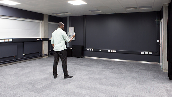 A black male stands alone in an empty room holding a remote device in his hand. In front of him is a black blank virtual reality wall. The digitalised data screen is wide.