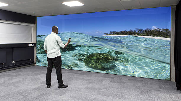 Virtual Reality Wall A black male stands alone in an empty room holding a remote device in his hand. A beautiful digitalised image of a large tropical seascape is displayed on a virtual reality wall in front of him. The digitalised data screen is huge covering the full size of the wall. large screen stock pictures, royalty-free photos & images