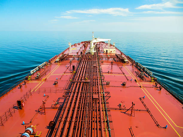 Deck of a crude oil carrier proceeding through blue ocean. Photo of a  big red oil tanker. There is long oil pipeline and cargo crane on deck of this ship. Vessel is steaming in blue, calm water to skyline under blue sky with smooth white clouds. oil tanker stock pictures, royalty-free photos & images