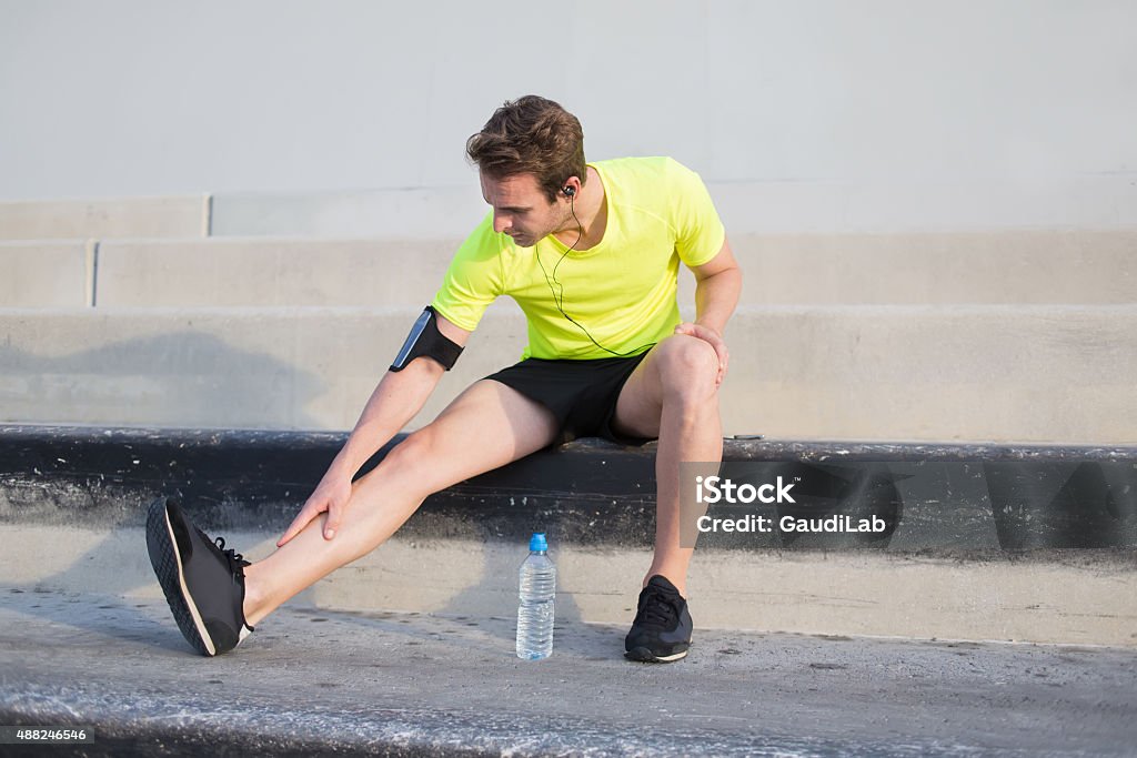 Men runner touching pulled leg muscle while resting after training Full length portrait of male runner touching pulled leg muscle as a result of fitness training outdoors, young sportsman taking break after get injured in the middle of morning running outside of city 2015 Stock Photo