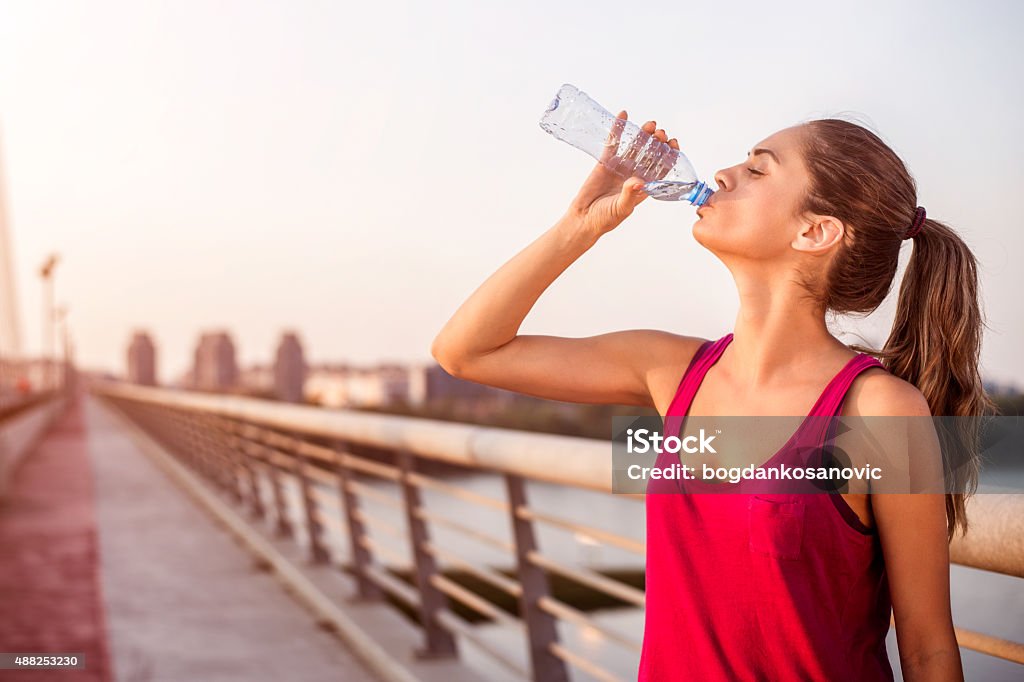 Sportswoman hydrating on the bridge Young sportswoman with pony tail drinking water on the bridge while resting from morning jogging. Drinking Stock Photo