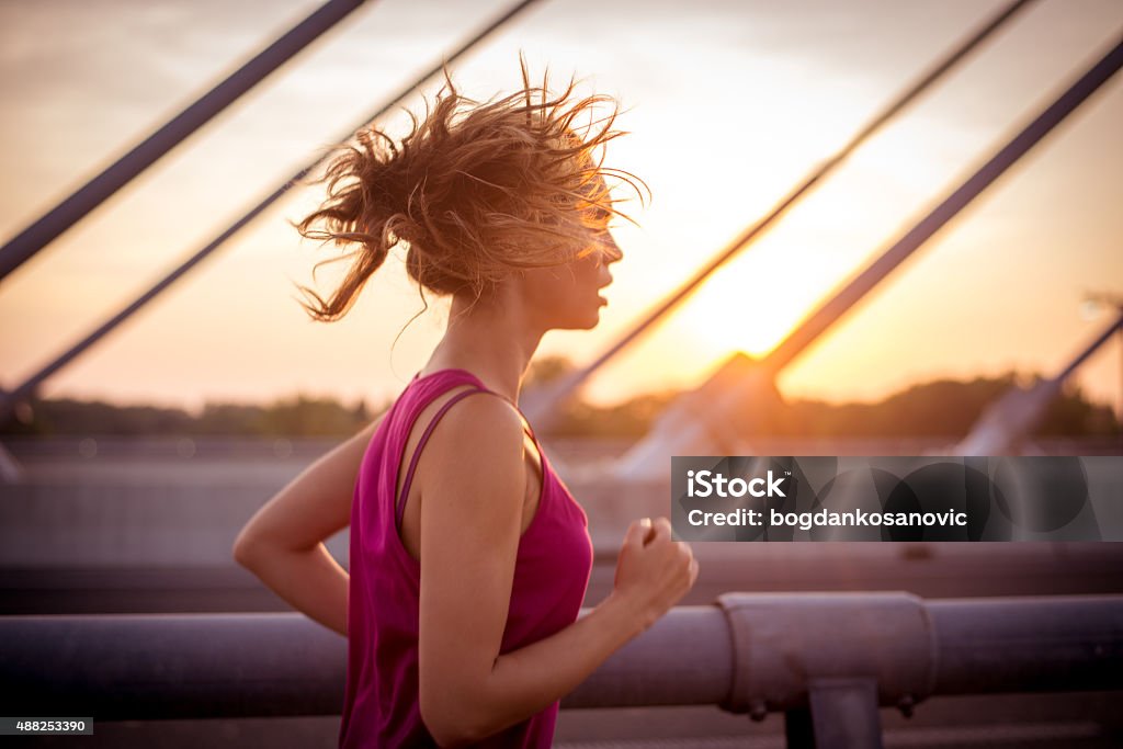 Female athlete running over the bridge in the morning Young sportswoman jogging over the bridge in the morning while her long brown hair is waving in the wind. Rising sun is in the background. Running Stock Photo