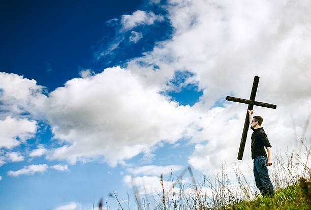 A man stands on a hilltop lifting up a large wooden cross toward the sky.  A depiction of a Christians victory in Jesus Christ over death through his resurrection as celebrated on Easter.  Horizontal image with copy space in the clouds.
