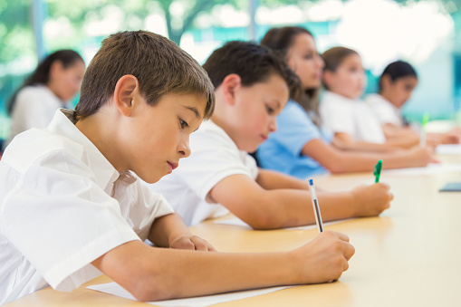Elementary age Hispanic little boy is sitting at desk with classmates in private elementary school. He is using a pen to write on paper during exam. Classmates are also concentrating while taking test. Students are wearing white and blue school uniforms.