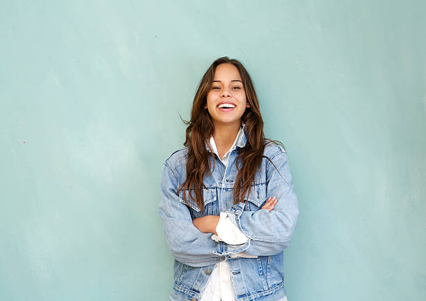 Young woman laughing with arms crossed against blue background Horizontal portrait of a young woman laughing with arms crossed in relaxed pose against blue background denim jacket stock pictures, royalty-free photos & images