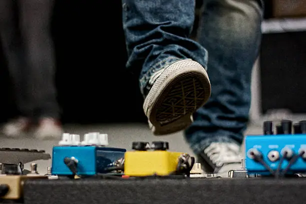 A close up of a guitarist’s foot midair about to step on a guitar pedal on stage at an outdoor venue.