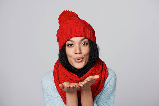 Christmas Girl. Mixed race african american - caucasian woman wearing knitted warm scarf and hat blowing a kiss, looking at camera, over gray background