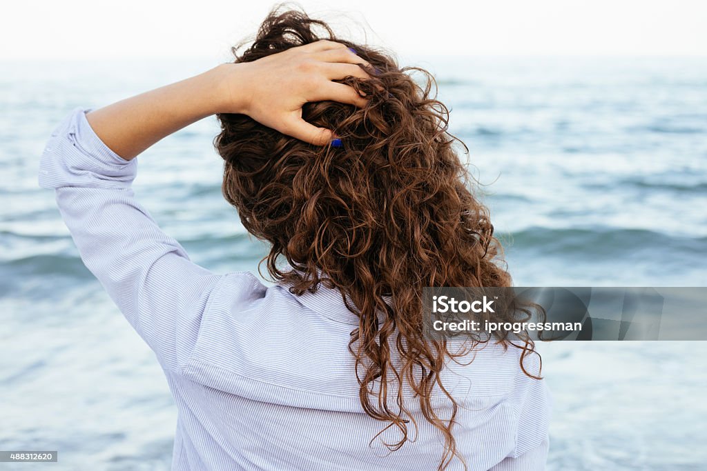 Young woman in shirt looking at the sea Young woman in shirt looking at the sea and keeps her hair. Back view close-up. Curly Hair Stock Photo