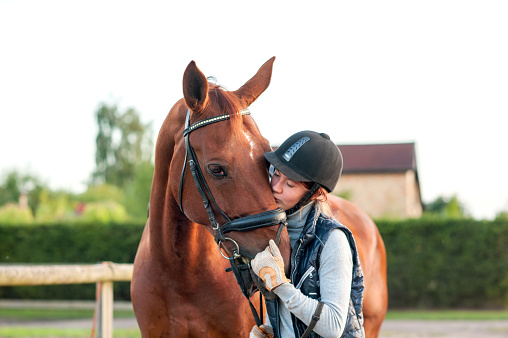 Young teenage girl equestrian kissing her chestnut horse. Multicolored outdoors horizontal image.