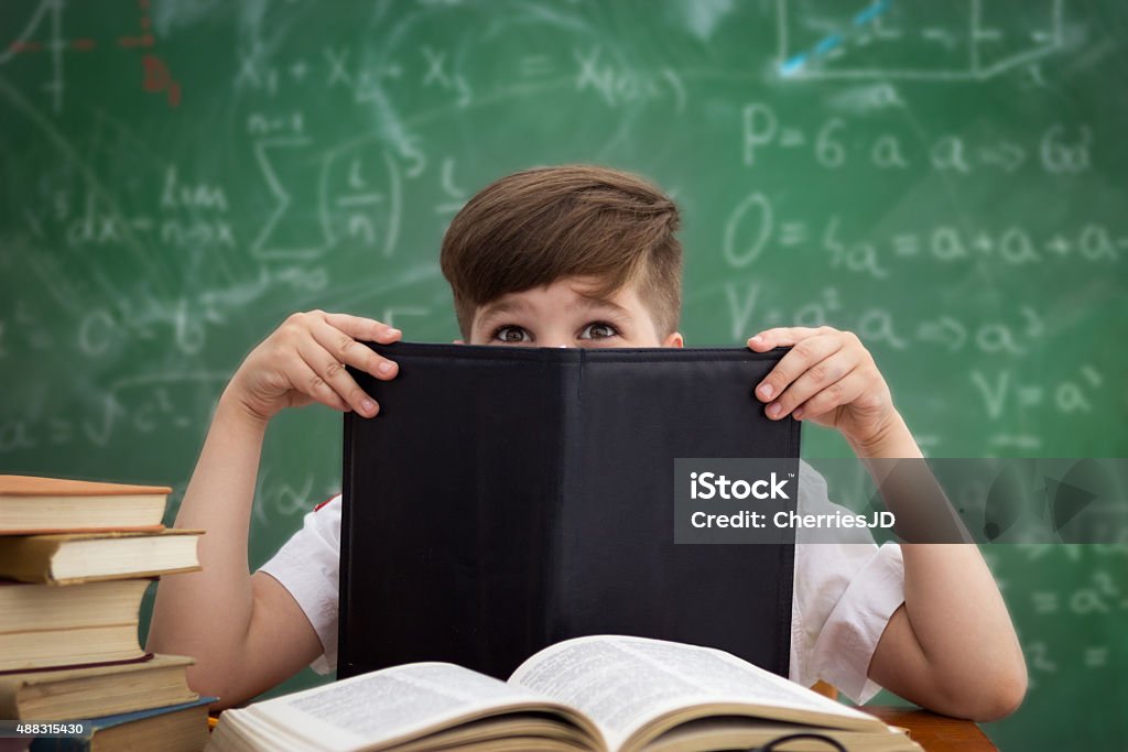 Funny schoolboy hiding behind book Funny schoolboy hiding behind book sitting in a classroom over a blackboard 2015 Stock Photo