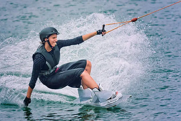 Woman touching the water with one hand and holding a rope with the other hand while wake boarding on dark-blue water.  She is wearing a black helmet and black wet suit.