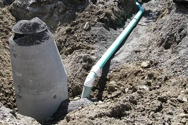 A catch basin storm drain attached to the main storm sewer pipe. A precast concrete manhole chamber is located adjacent to the drain pipe, granular material rests on the frame and cover of the manhole.