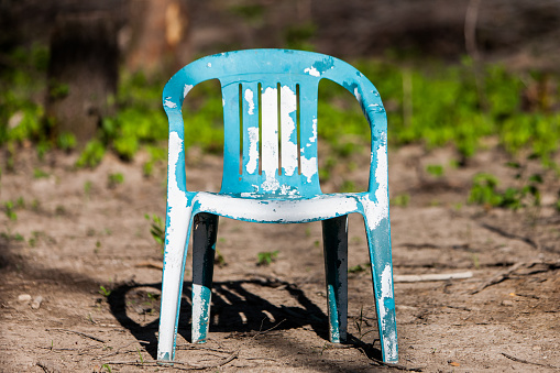 A horizontal image shows view of a painted blue chair with chipping paint. The chair sits on a dirt ground outside.