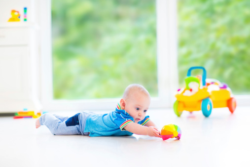 Adorable baby boy playing with a colorful ball and toy car in a sunny nursery with white furniture and white floor and a big garden view window