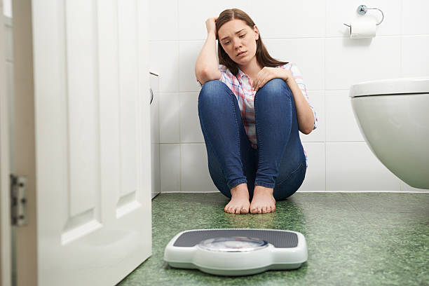 Unhappy Teenage Girl Sitting On Floor Looking At Bathroom Scales stock photo