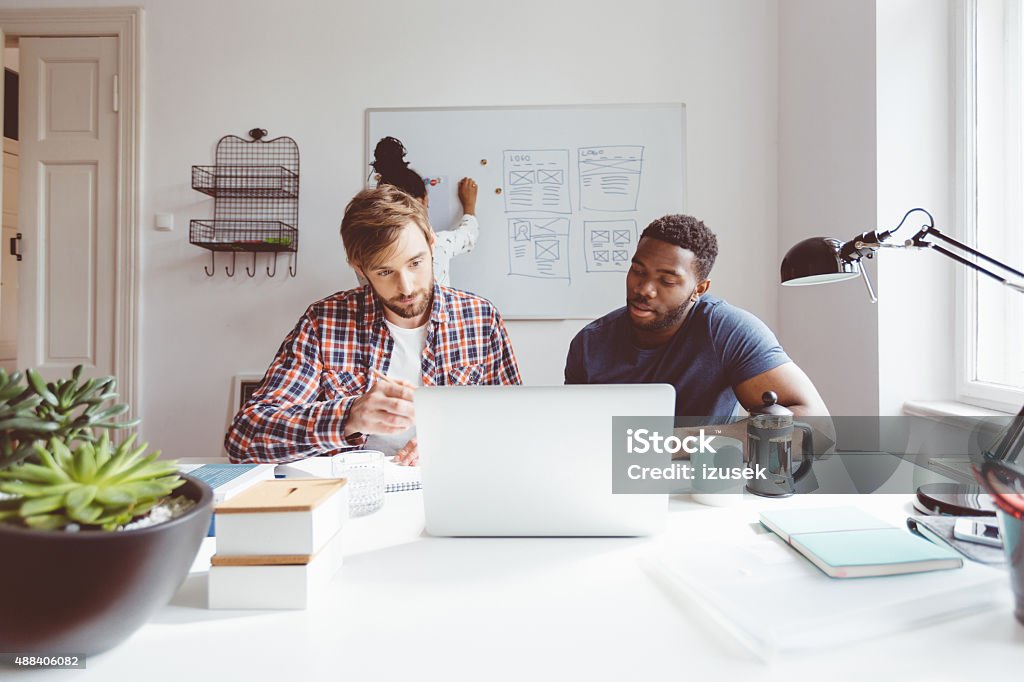 Bearded blonde man working on laptop with afro american colleague Start-up or advertising agency. Bearded blonde man and afro american man brainstorming in an office, using laptop together.  Computer Stock Photo