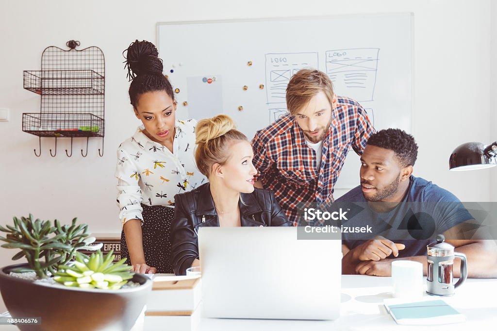 Start-up agency, multi ethnic team brainstorming, using laptop Start-up or advertising agency. Multi ethnic team of designers - caucasian and afro american - meeting in an office, sitting at the table, discussing, brainstorming, using laptop. Persuasion Stock Photo