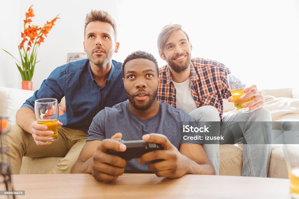 Three guys playing video games Multi ethnic - caucasian and afro american - friends sitting on sofa at home, playing video games, drinking beer. Close up of faces. Video Game Stock Photo