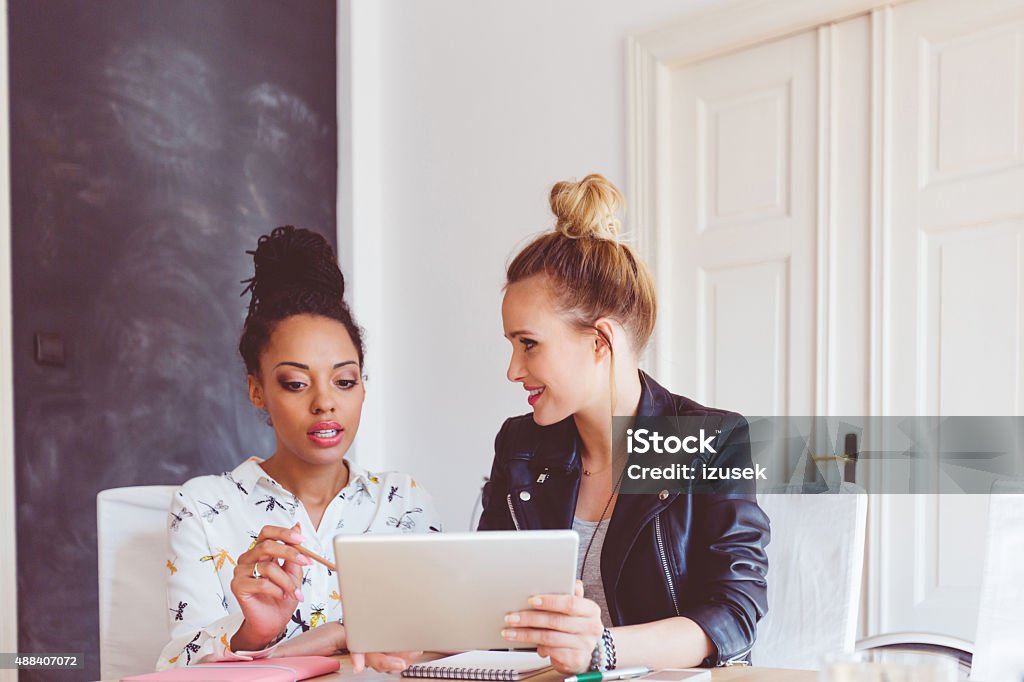 Two women working on digital tablet in an office Start-up or advertising agency. Two women - caucasian and afro american - sitting at the table in an office and using a digital tablet together.   Social Media Stock Photo