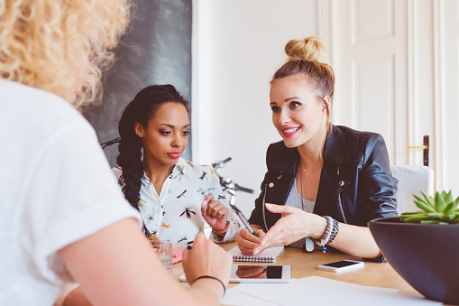 Start-up or advertising agency. Three women - caucasian and afro american - sitting at the table in an office and discussing. Digital tablet and smart phone ont he table.