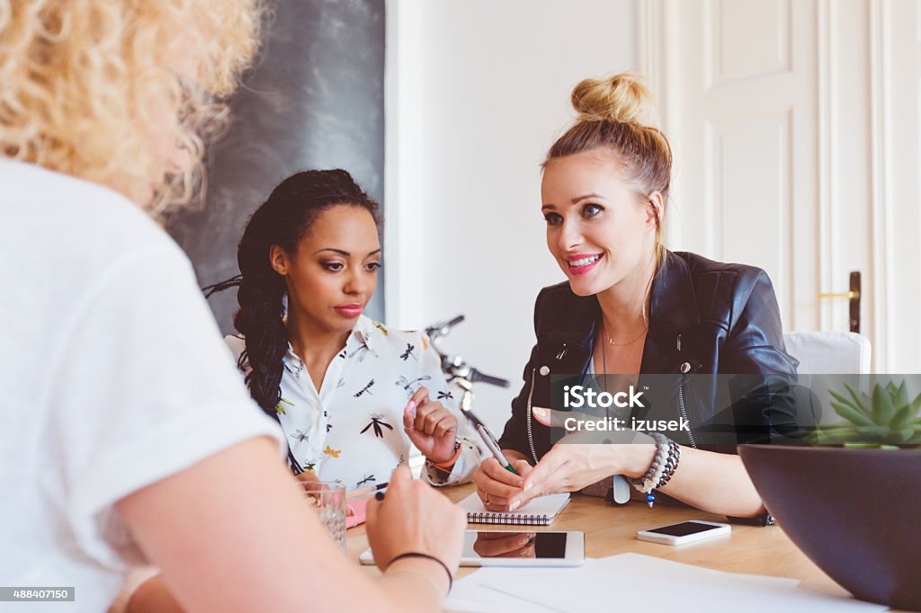 Three women discussing in an office Start-up or advertising agency. Three women - caucasian and afro american - sitting at the table in an office and discussing. Digital tablet and smart phone ont he table. Persuasion Stock Photo