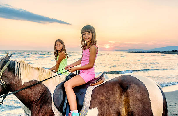 due sorelle cavalcare cavalli nel tramonto in riva al mare - foal child mare horse foto e immagini stock