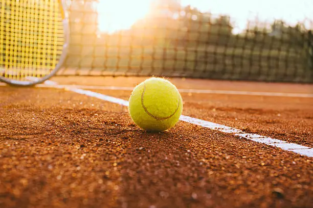 Tennis racket and ball on a clay court with sun in backlight.
