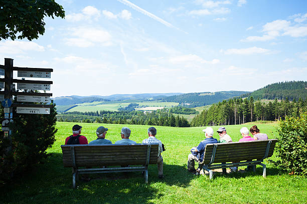 Langewiese, Germany - August 22, 2015: Summershot of seniors having a break on benches at trekking pathway Rothaarsteig in Sauerland. Scene is in Langewiese, small village near to Neuastenberg and Winterberg. Between seniors is an adult woman. Gorup is watching to north.
