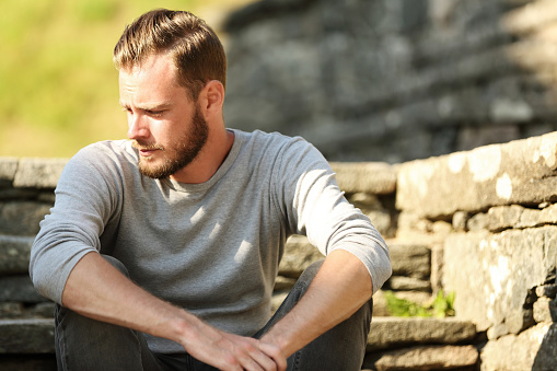 Man in his 20s wearing a grey shirt and jeans, sitting down outside on a set of steps on a sunny summer day. 