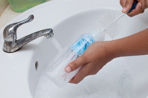 Mother hand washing baby milk bottle on white sink and water drop from faucet