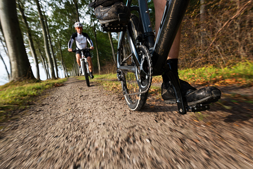 Dramatic low angle view showing  a male mountain biker following another cyclist along a forest track next to the sea . Taken in Fanefjord Woods on the island of Møn in Denmark. The picture is cropped so the head of the rider in front head is not shown in the picture, with the rider following out of focus in the background. Shallow focus point on the front chain ring of the bike. Horizontal format with some copy space.