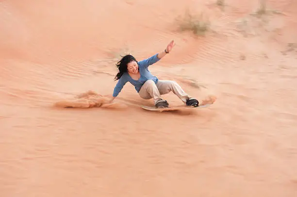 Dubai iStockalypse.  A happy, mature, Asian woman solo adventure tourist loses balance and is about to crash on her sandboard at the bottom of a sand dune in the red desert between Dubai and Sharjah, United Arab Emirates.  Closeup view with copyspace.
