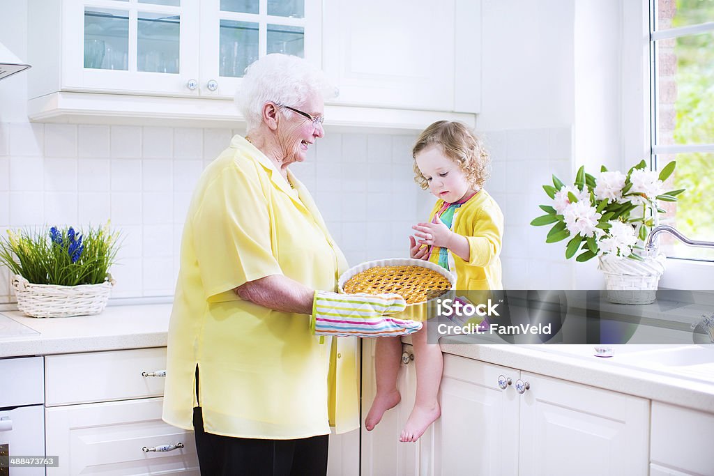 Sweet granddaughter with her pretty great grandmother baking apple pie Happy beautiful great grandmother and her adorable granddaughter, curly toddler girl in colorful dress, baking an apple pie together standing next to white oven in sunny modern kitchen with big window Active Seniors Stock Photo