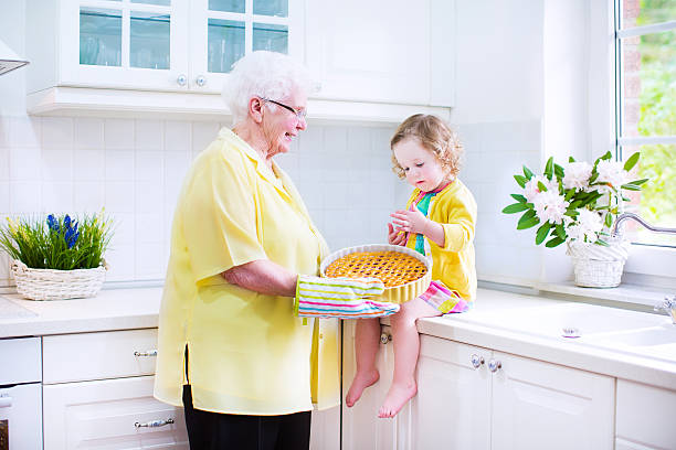 Sweet granddaughter with her pretty great grandmother baking apple pie Happy beautiful great grandmother and her adorable granddaughter, curly toddler girl in colorful dress, baking an apple pie together standing next to white oven in sunny modern kitchen with big window fume cupboard stock pictures, royalty-free photos & images