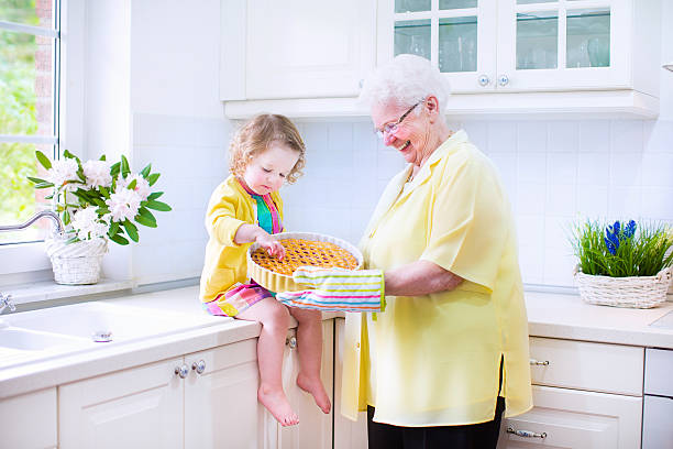 Little granddaughter with her pretty great grandmother baking apple pie Happy beautiful great grandmother and her adorable granddaughter, curly toddler girl in colorful dress, baking an apple pie together standing next to white oven in sunny modern kitchen with big window fume cupboard stock pictures, royalty-free photos & images