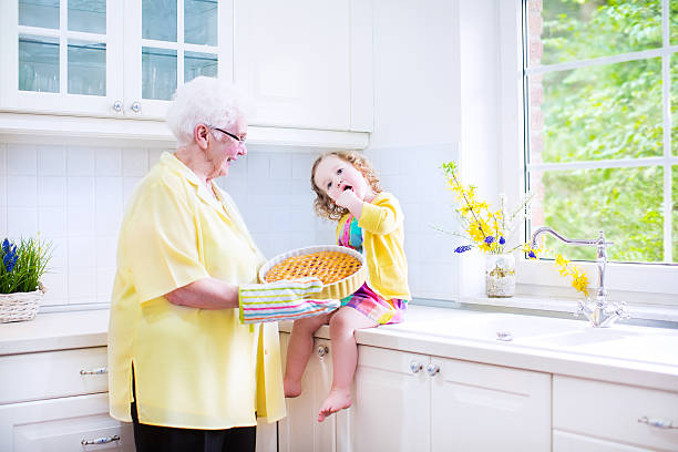 Great grandmother and little girl cooking pie in white kitchen Happy beautiful great grandmother and her adorable granddaughter, curly toddler girl in colorful dress, baking an apple pie together standing next to white oven in sunny modern kitchen with big window fume cupboard stock pictures, royalty-free photos & images