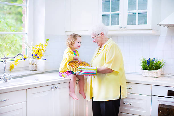 Great grandmother with her granddaughter cooking pie in white kitchen Happy beautiful great grandmother and her adorable granddaughter, curly toddler girl in colorful dress, baking an apple pie together standing next to white oven in sunny modern kitchen with big window fume cupboard stock pictures, royalty-free photos & images