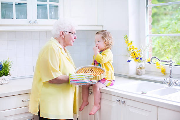 Adorable grandmother and sweet girl baking pie in white kitchen Happy beautiful great grandmother and her adorable granddaughter, curly toddler girl in colorful dress, baking an apple pie together standing next to white oven in sunny modern kitchen with big window fume cupboard stock pictures, royalty-free photos & images