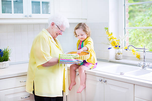 Happy grandmother, granddaughter standing next white oven in sunny kitchen Happy beautiful great grandmother and her adorable granddaughter, curly toddler girl in colorful dress, baking an apple pie together standing next to white oven in sunny modern kitchen with big window fume cupboard stock pictures, royalty-free photos & images