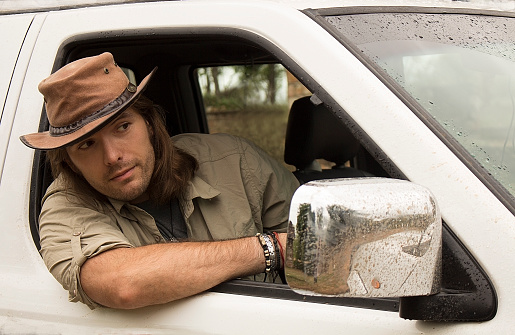 Handsome young man with long hair in beige brown cowboy hat driving white off road 4x4 car after rain. Safari style. 