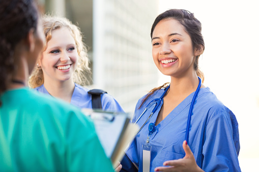 Young adult Hispanic woman is talking with young adult Caucasian blonde woman and young adult African American woman outside on college campus. Women are nursing or medical students. They are wearing hospital scrubs and stethoscopes.
