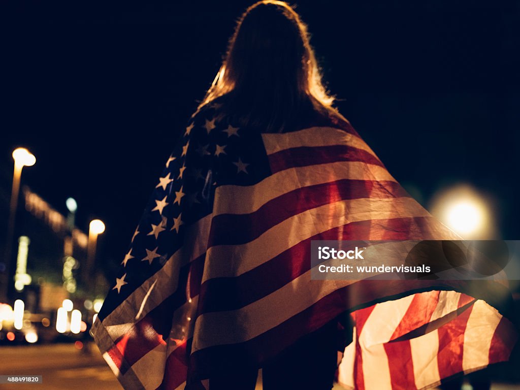 Woman draped with American flag on city street at night Rearview of a young woman with an American flag draped over her shoulders walking alone on city street at night with street lights shining through the shadows of the flag American Flag Stock Photo