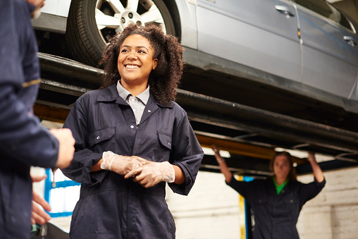 A young female adult mechanic is chatting next to a car on a ramp . She is wearing blue overalls and protective gloves  . In the background a car is seen on a hydraulic lift with a  female mechanic underneath it .