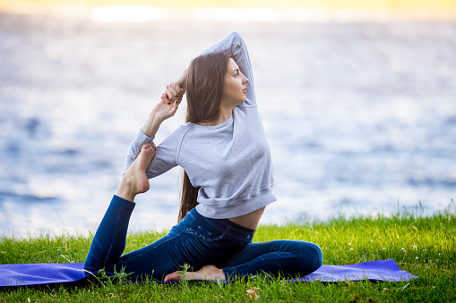 Profile of beautiful sporty young woman working out on city street, doing warming up stretching exercises on lake in park, sitting in Eka Pada Rajakapotasana, full length